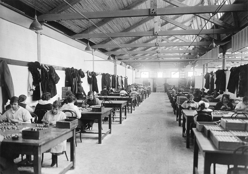 Vintage black and white photograph of a factory interior with workers at desks and hanging clothing.